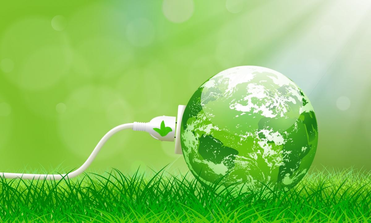 Een groene wereldbol die via een witte stekker is aangesloten op stroom, liggend in het gras tegen een zachte, groen getinte achtergrond als symbool voor duurzame energie.