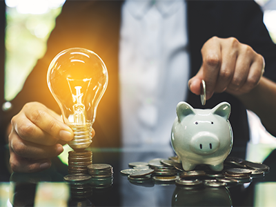 Persoon plaatst een munt in een spaarvarken terwijl hij een brandende gloeilamp vasthoudt, omgeven door stapels munten – symbool voor energiebesparing en financiële winst.