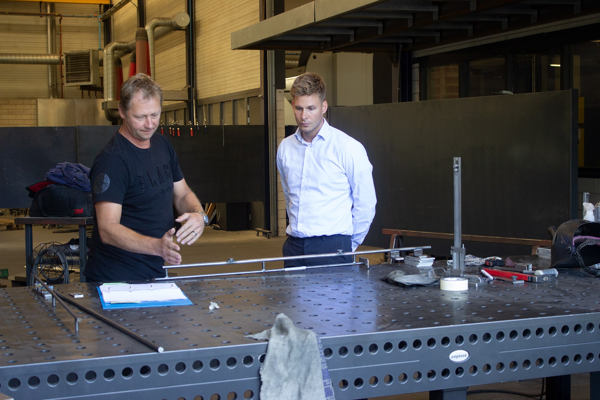 Twee mannen in een werkplaats bespreken een technische tekening aan een metalen werktafel, omringd door gereedschap.