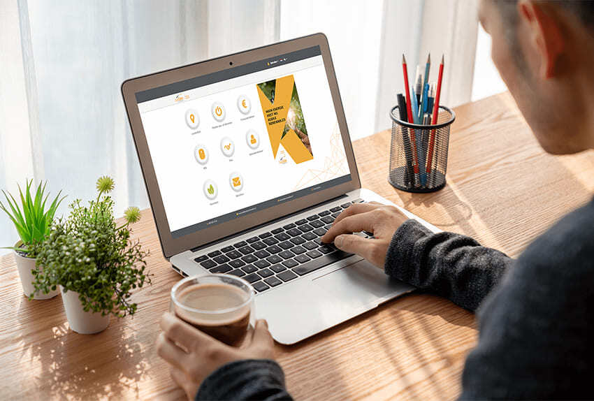 Man werkt aan een houten bureau met een laptop waarop het portaal 'Mijn Audax Renewables' geopend is, met een kop koffie in de hand en omringd door groene planten en een pot met pennen.