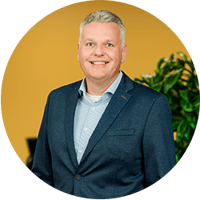 Een man met kort grijs haar in een donkerblauw colbert en gestreept overhemd, lachend voor een gele achtergrond met een groene plant op de achtergrond.