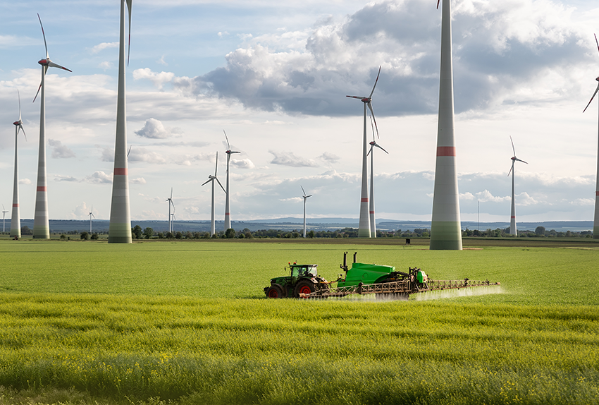 Tractor besproeit een groen landbouwveld met op de achtergrond meerdere windmolens die duurzame energie opwekken onder een deels bewolkte hemel.