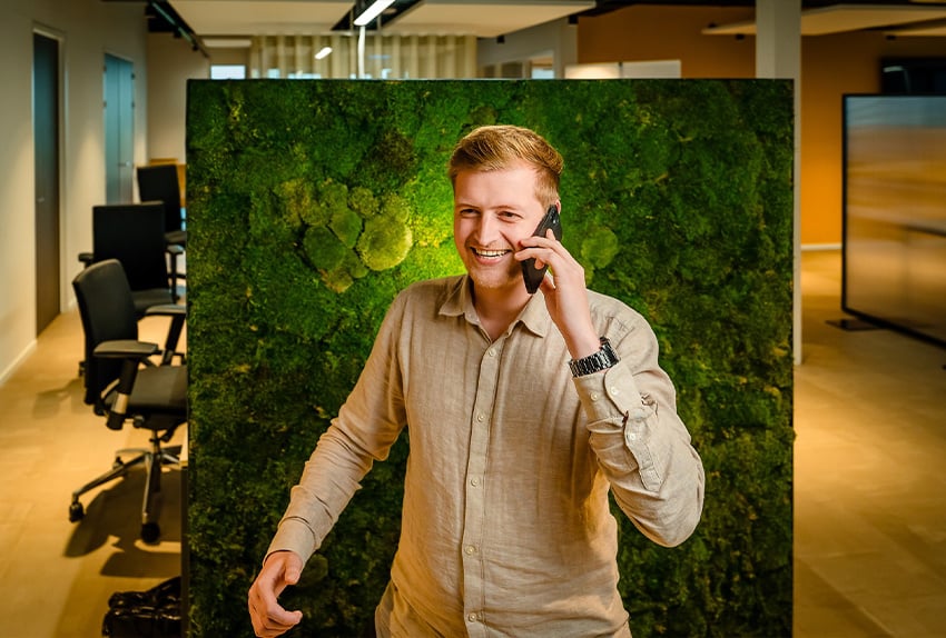 Lachende man in een kantooromgeving voert een telefoongesprek, staand voor een groene moswand.