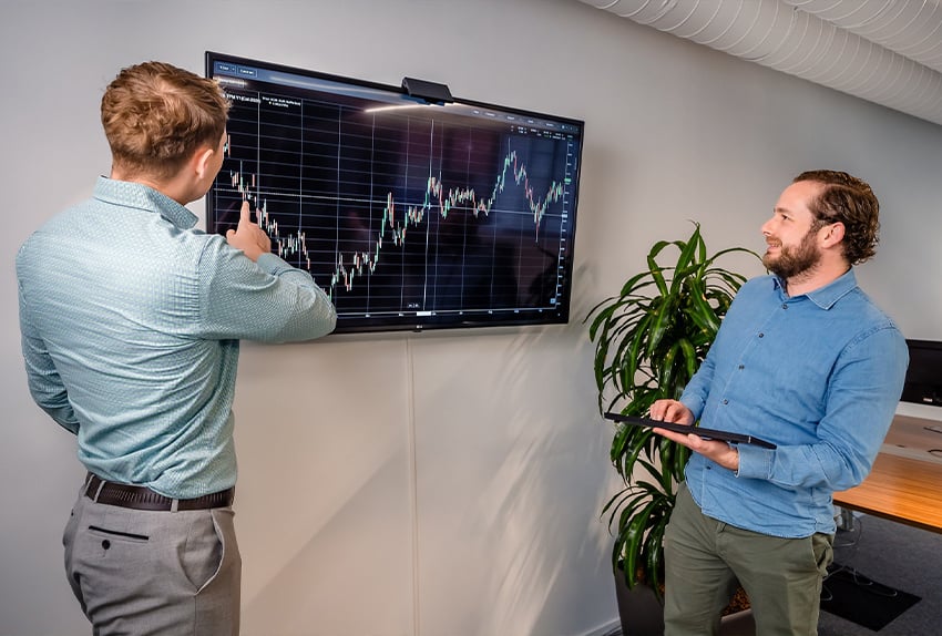 Twee mannen analyseren een grafiek op een groot scherm in een moderne kantoorruimte.