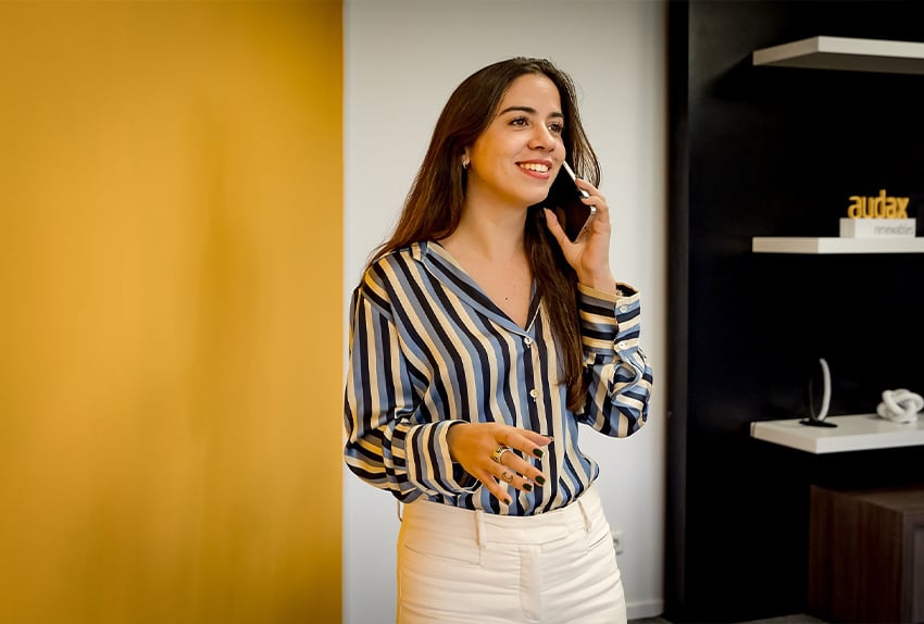 Vrouw met lang bruin haar en gestreepte blouse voert een telefoongesprek en glimlacht in een moderne kantooromgeving met geel-witte en zwarte accenten.