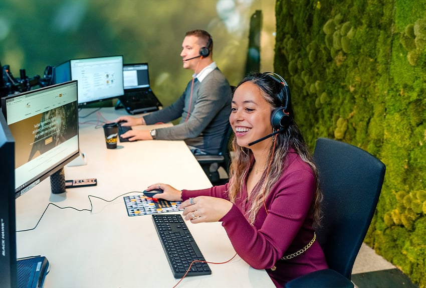 Twee klantenservicemedewerkers met headset werken lachend aan hun bureau bij Audax Renewables, met groene moswand op de achtergrond.
