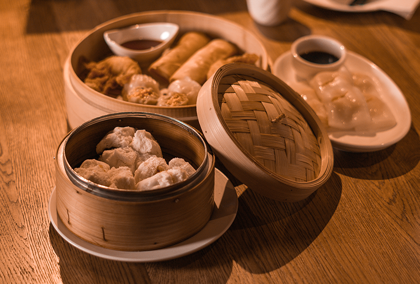 Assortiment Aziatische dim sum-gerechten geserveerd in traditionele bamboe stoommandjes op een houten tafel, vergezeld van kleine schaaltjes met dipsauzen.