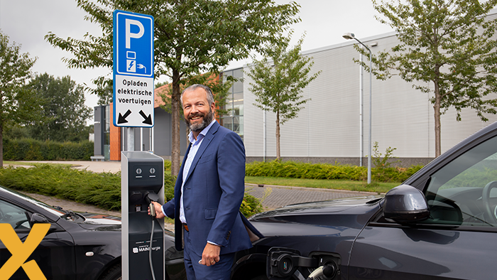Zakenman in pak laadt een elektrische auto op bij een laadpaal op een parkeerplaats met bord voor opladen van elektrische voertuigen.