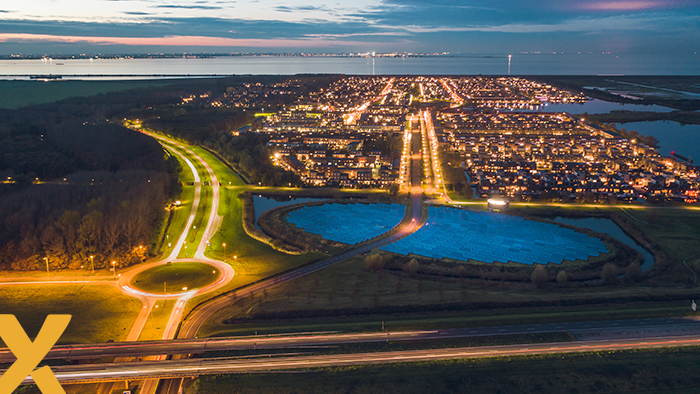 Luchtfoto van een verlichte stad bij schemering met zonnepark en snelweg op de voorgrond.