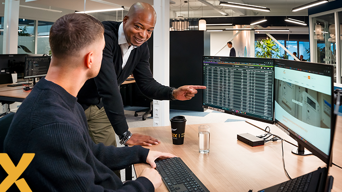 Twee collega’s in een modern kantoor kijken naar data op computerschermen, terwijl één persoon iets aanwijst.