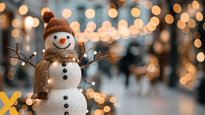 Sneeuwpop met sjaal en muts, versierd met lichtjes, in een sfeervol verlichte straat met kerstverlichting op de achtergrond.