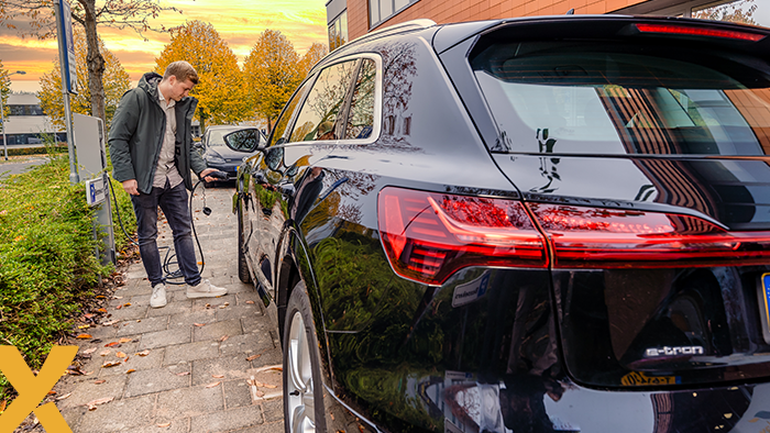 Man sluit een elektrische auto aan op een laadpaal langs de stoep in een woonwijk tijdens zonsondergang.