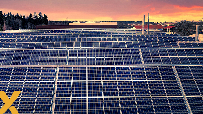 Dak van een bedrijfspand volledig bedekt met zonnepanelen bij zonsondergang, met fabriekspijpen op de achtergrond.