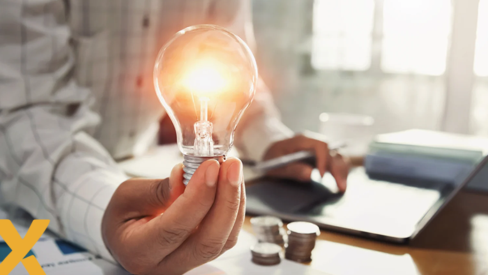 Hand houdt een brandende gloeilamp vast voor een laptop en stapel munten, symbool voor slimme energiebesparing en kostenbeheer.