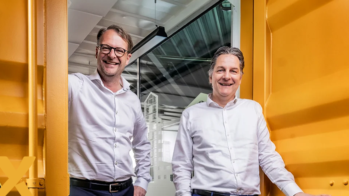 Twee lachende mannen in witte overhemden poseren in een moderne kantoorruimte met gele accenten.