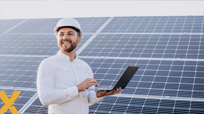 Lachende man met veiligheidshelm en laptop voor een veld met zonnepanelen, symbool voor monitoring en beheer van zonne-energie.