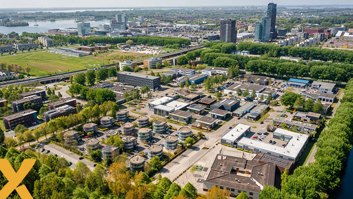 Luchtfoto van een groen bedrijventerrein aan de rand van een stad met hoogbouw en water op de achtergrond.