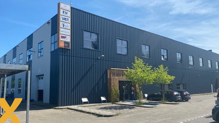 Zwartgekleurd bedrijfsgebouw met gevelborden, parkeerplaatsen met enkele auto’s en een helderblauwe lucht erboven.
