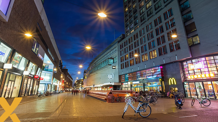 Verlichte winkelstraat bij avond met moderne winkelpuien, fietsenrekken en geparkeerde fietsen, met mensen op de achtergrond.