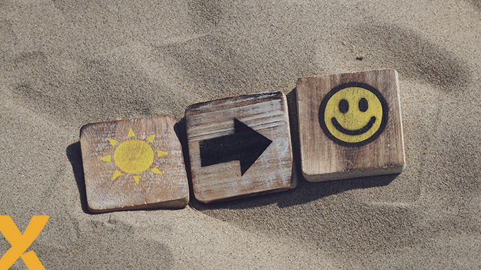 Drie houten blokjes in het zand met symbolen van een zon, een pijl en een smiley, die een positieve richting aangeven.