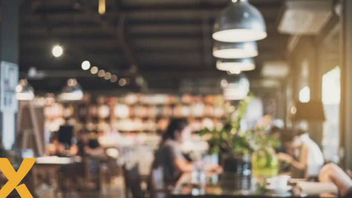 Wazige achtergrond van een gezellig ingericht café of restaurant met mensen aan tafels, sfeervolle verlichting en een groene plant op de voorgrond.