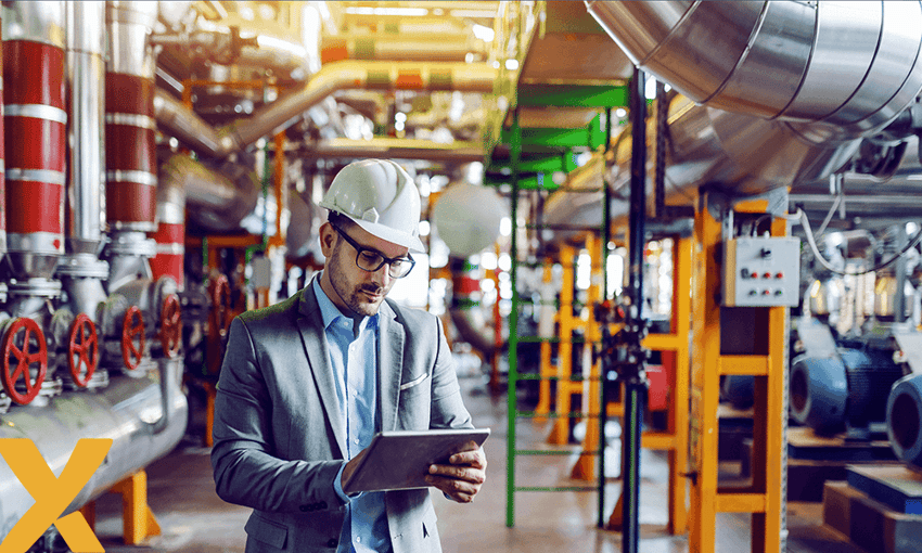 Een man in pak en witte veiligheidshelm die in een industriële fabriekshal gegevens op een tablet bekijkt.