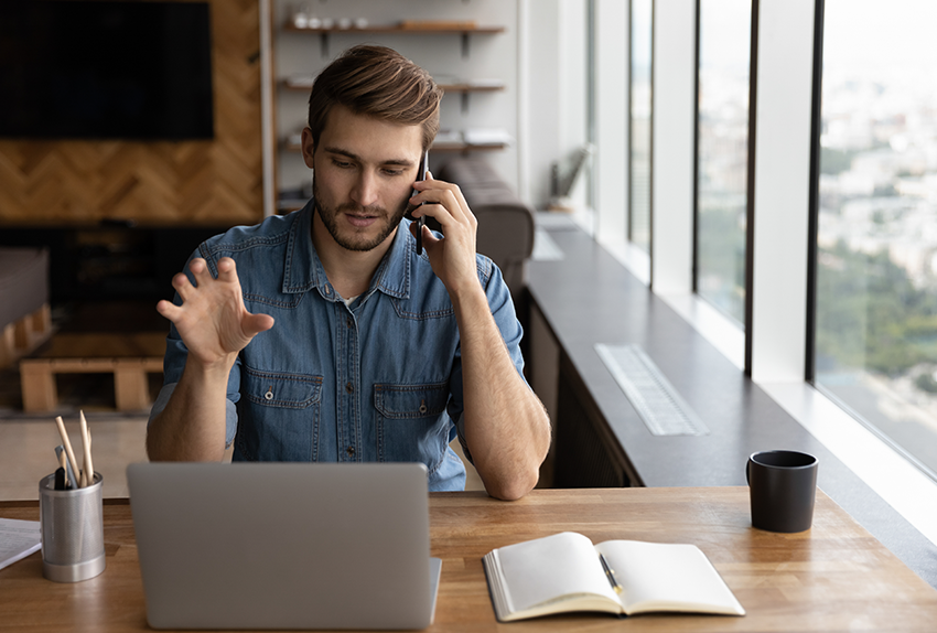Man voert een telefoongesprek terwijl hij thuis werkt aan een houten bureau met laptop, notitieboek en koffiebeker, met uitzicht op de stad door grote ramen.
