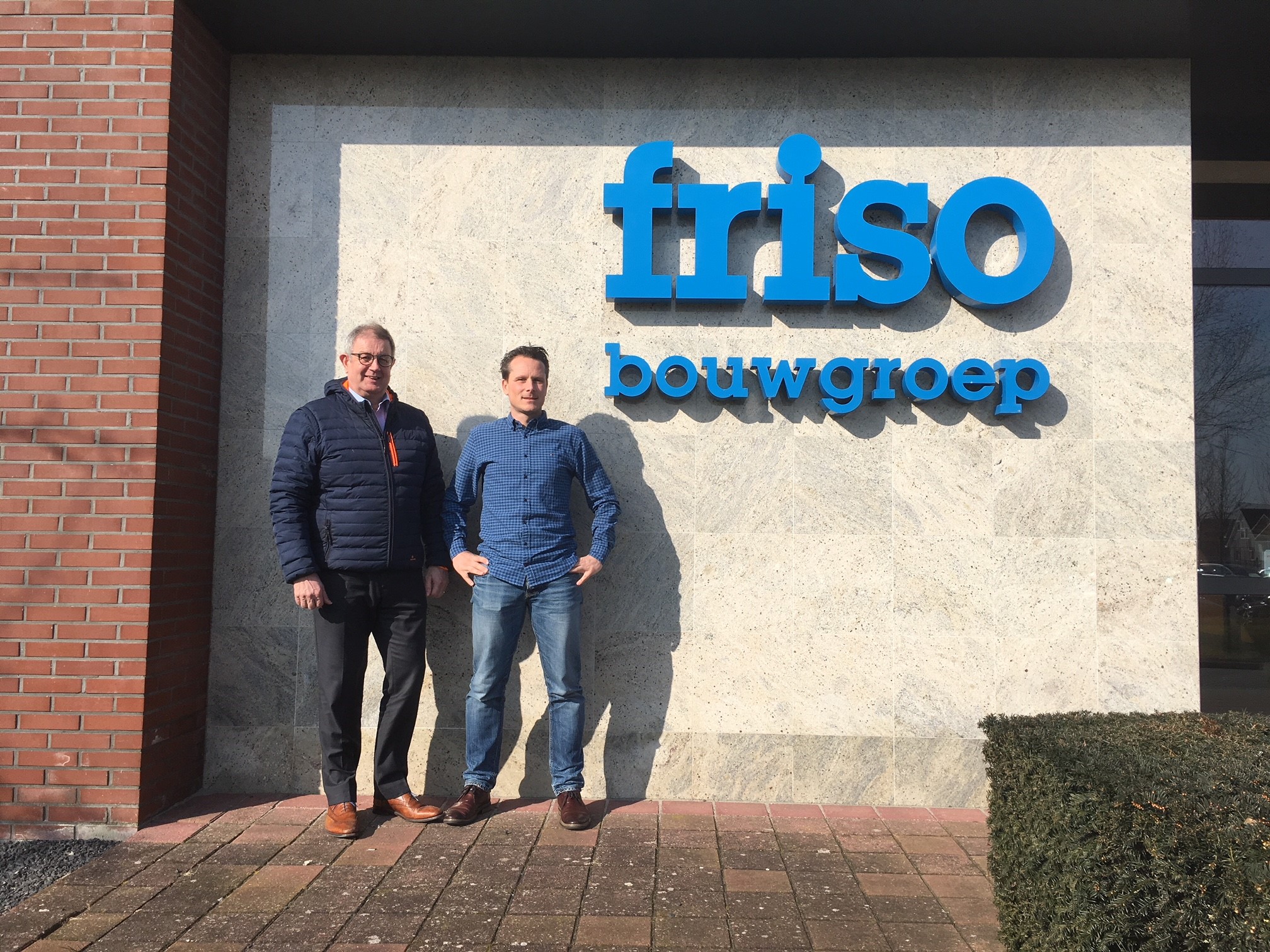 Twee mannen poseren voor het logo van Friso Bouwgroep op een gevelmuur, onder een zonnige hemel.