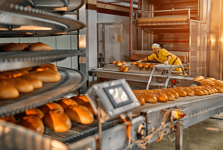 Een bakker werkt in een industriële bakkerij en plaatst versgebakken broden op een lopende band in een geautomatiseerd productieproces.