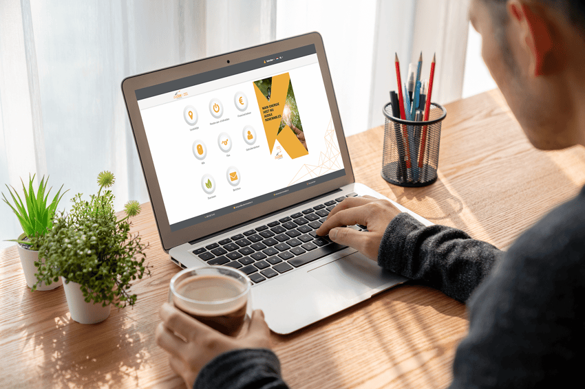 Man werkt aan een houten bureau met een laptop waarop het portaal 'Mijn Audax Renewables' geopend is, met een kop koffie in de hand en omringd door groene planten en een pot met pennen.