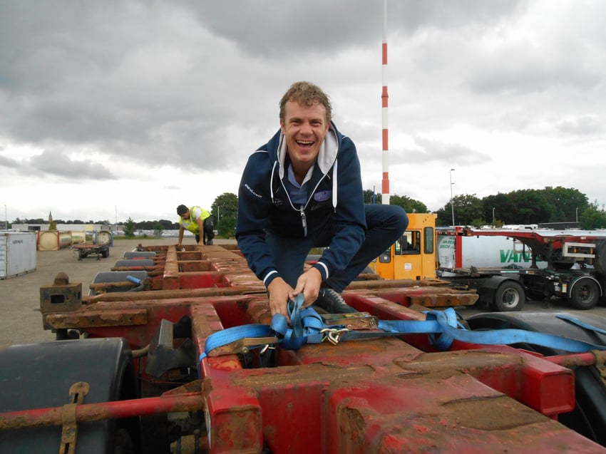 Lachende man bevestigt een spanband op een oplegger terwijl een collega op de achtergrond aan het werk is op een industrieterrein.