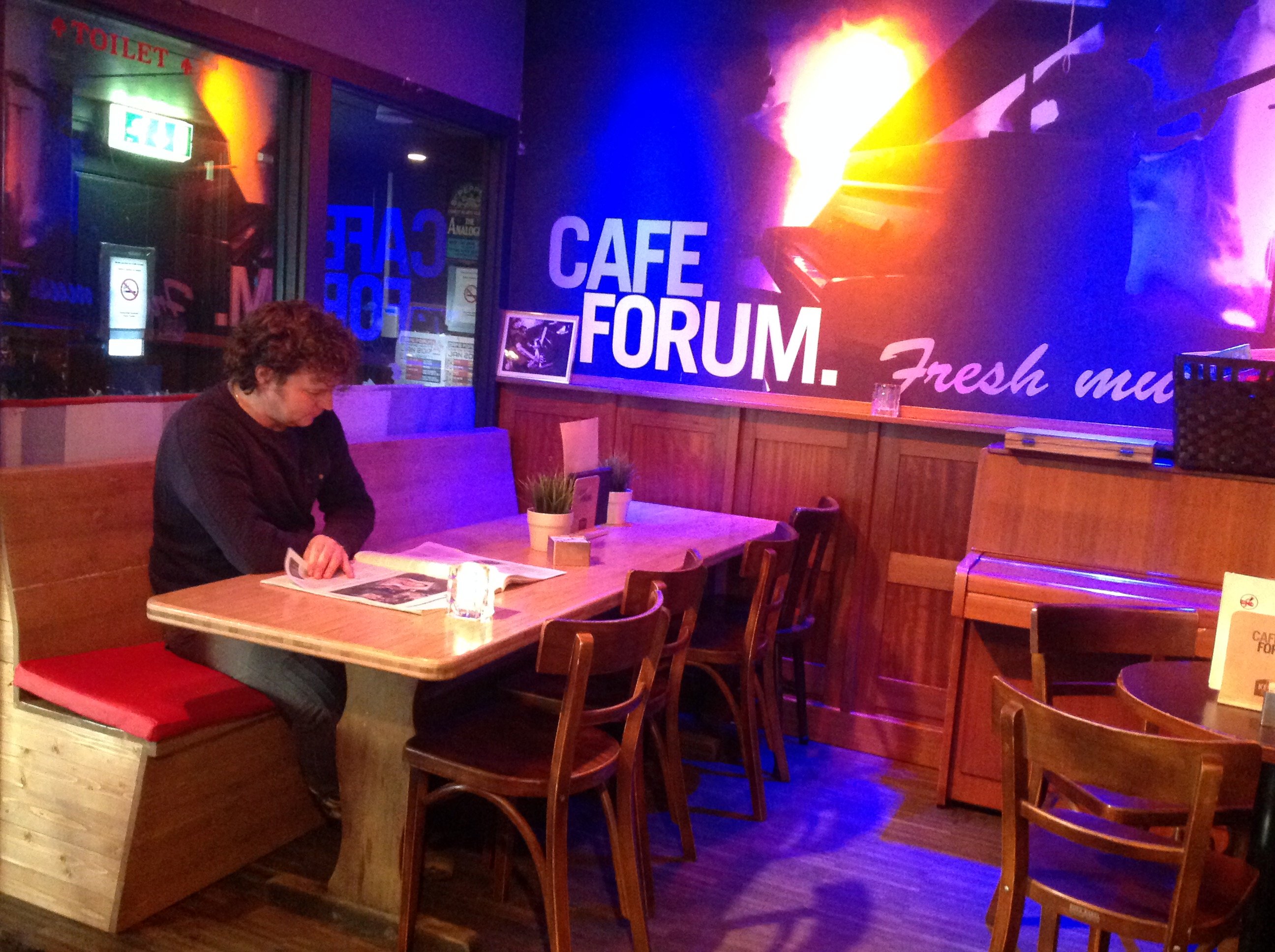 Man leest een tijdschrift aan een houten tafel in Café Forum, met sfeervolle verlichting en een muurdecoratie waarop 'CAFE FORUM. Fresh music' staat.