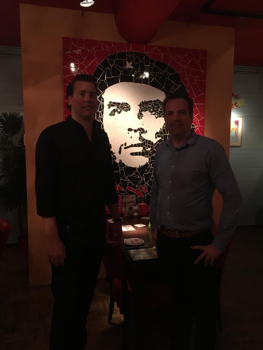 Twee mannen poseren in het sfeervolle restaurant van Cubanita Maastricht, voor een opvallend mozaïekportret van Che Guevara aan de muur.