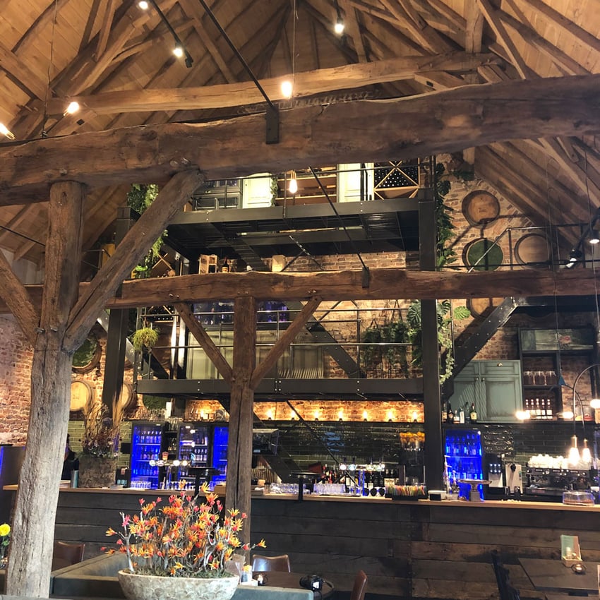 Sfeervol restaurantinterieur met hoge houten balken, een open mezzanine, blauw verlichte bar en industriële accenten.