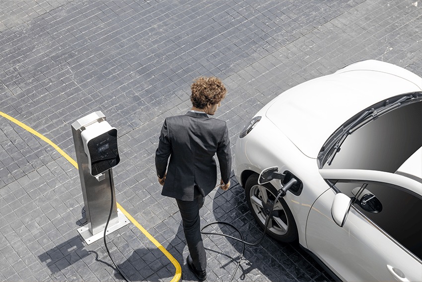 Man in pak loopt langs een witte elektrische auto die is aangesloten op een laadpaal op een betegeld parkeerterrein.