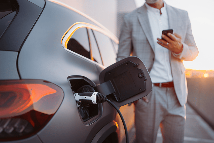 Elektrische auto wordt opgeladen terwijl een man in een grijs pak op zijn smartphone kijkt, bij zonsondergang op een parkeerplaats.