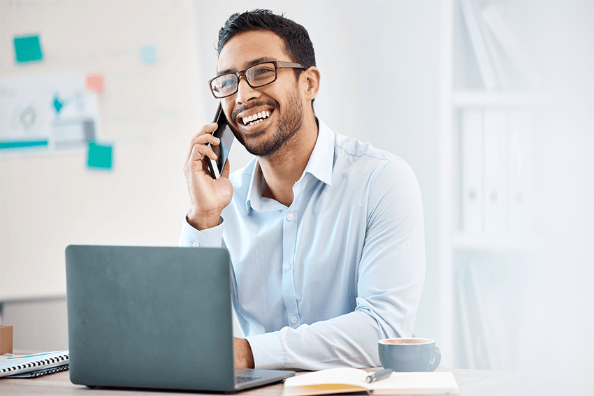 Vriendelijke kantoormedewerker met bril voert een telefoongesprek en lacht terwijl hij achter zijn laptop zit, met een kop koffie op het bureau.