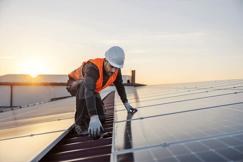Monteur met veiligheidshesje en helm installeert zonnepanelen op een dak bij zonsondergang, als onderdeel van een duurzaam energieproject.