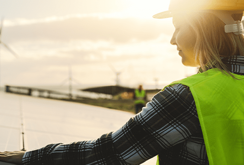 Vrouw in veiligheidsvest en helm bekijkt zonnepanelen op een zonnepark bij zonsondergang, met windmolens zichtbaar op de achtergrond.