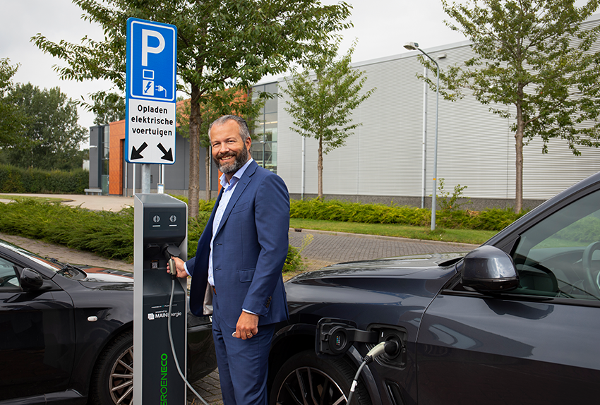 Man in pak laadt elektrische auto op bij een laadpaal op een bedrijventerrein, met een bord 'Opladen elektrische voertuigen' op de achtergrond.