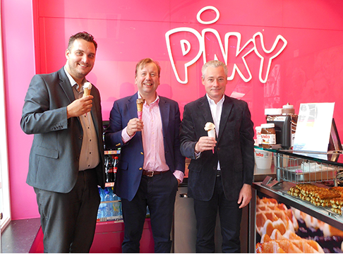 Drie mannen in pak genieten van een ijsje in een ijssalon met felroze muur en het logo 'PINKY' op de achtergrond.