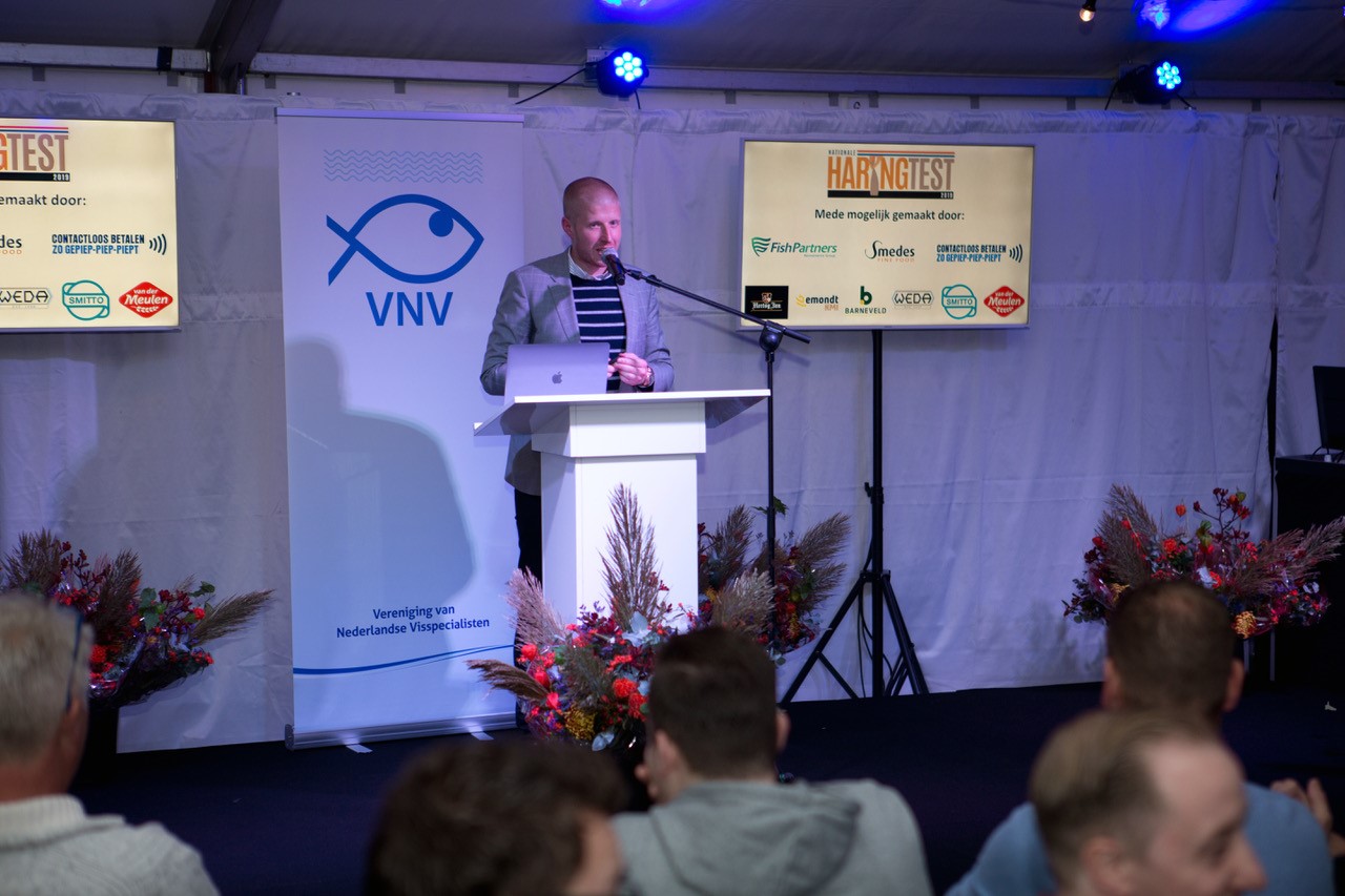Spreker bij een presentatie op een evenement van de Vereniging van Nederlandse Visspecialisten (VNV), met banners en schermen waarop sponsoren van de Haringtest worden getoond.
