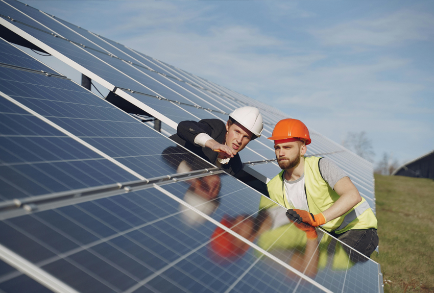 Twee mannen met veiligheidshelmen inspecteren zonnepanelen op een zonneveld onder een heldere hemel.