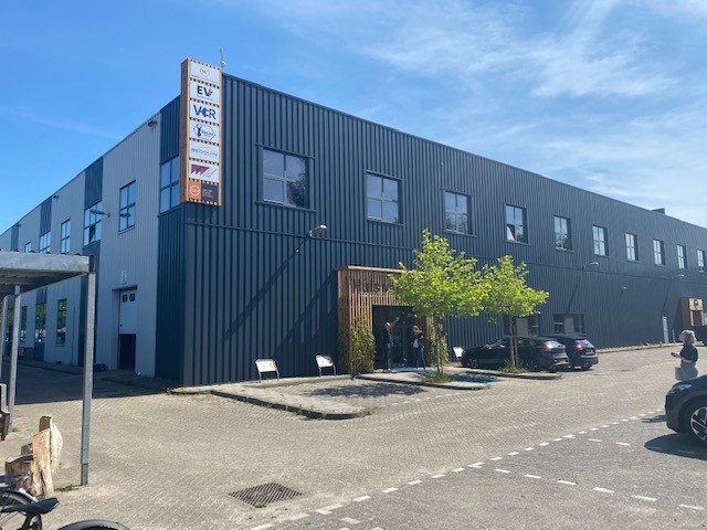 Zijkant van een modern industrieel bedrijfsverzamelgebouw met meerdere bedrijfsnaamborden aan de gevel, een entree met houten accent en enkele geparkeerde auto's onder een boom.