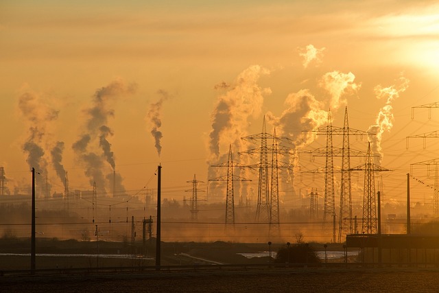 Industriële omgeving bij zonsondergang met meerdere rookpluimen uit fabrieksschoorstenen en hoogspanningsmasten op de voorgrond.